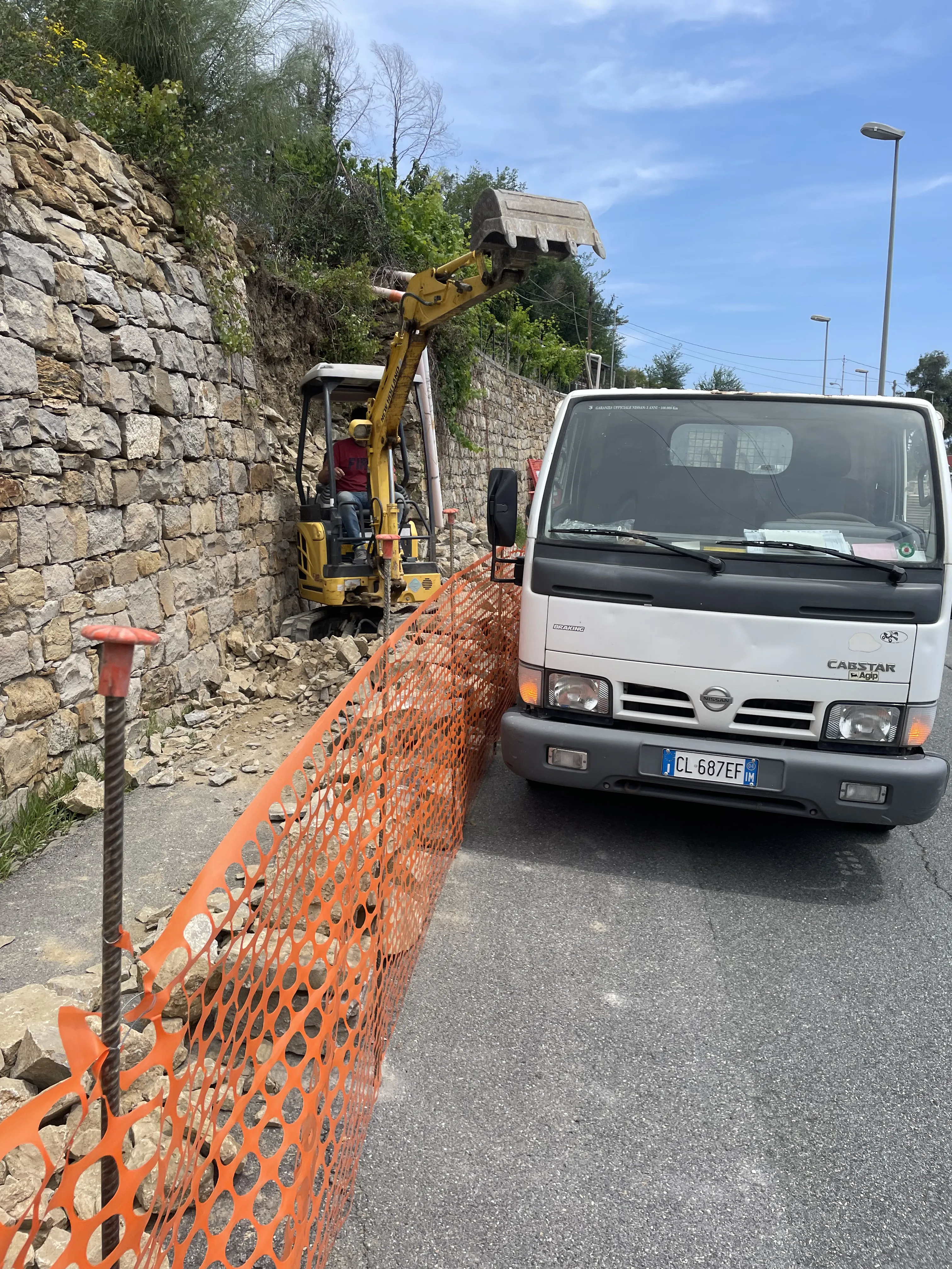 Mini escavatore in scavo accanto a muro in pietra, con camion